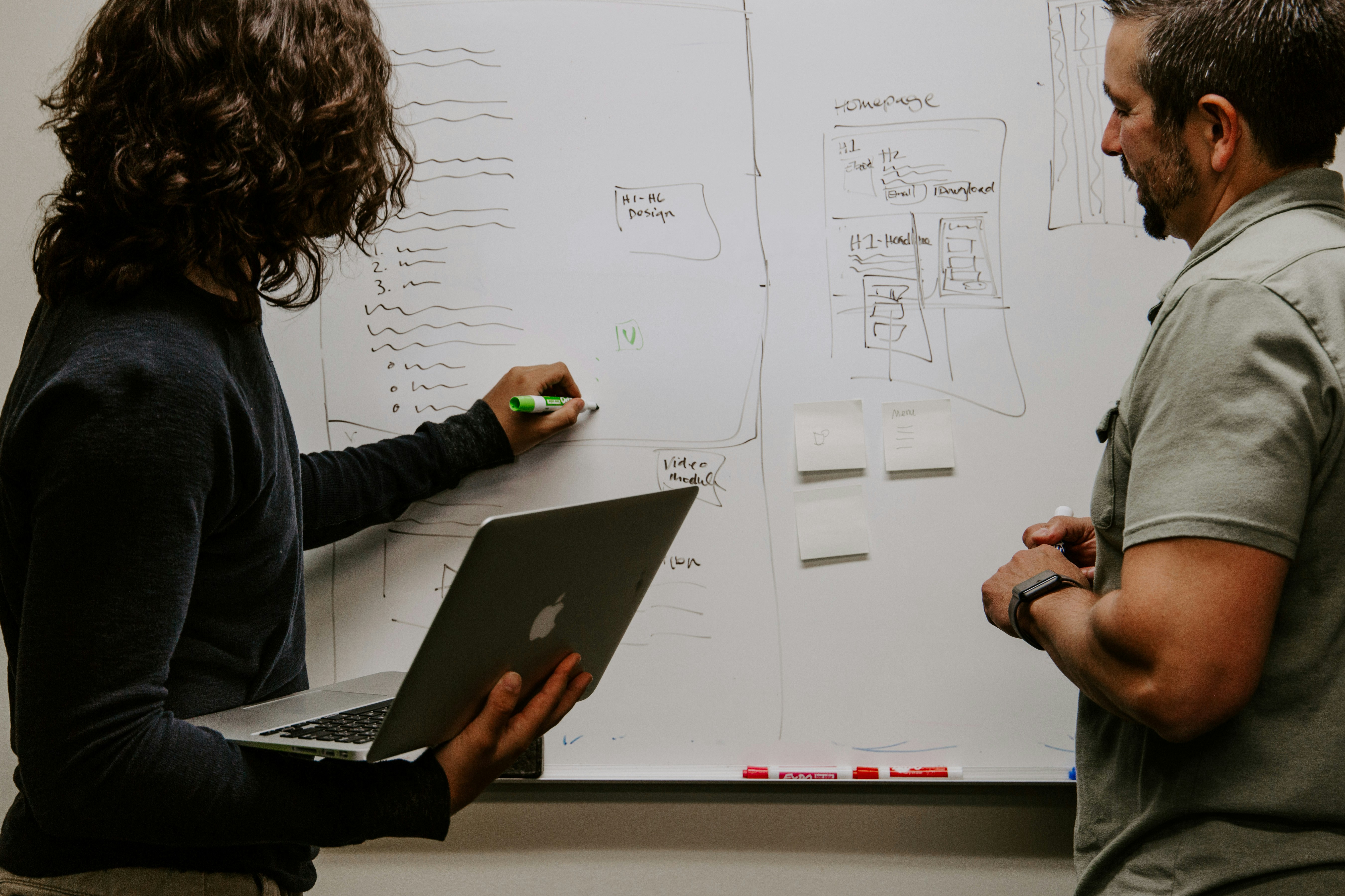 A woman with a computer and a man standing in front of a white board